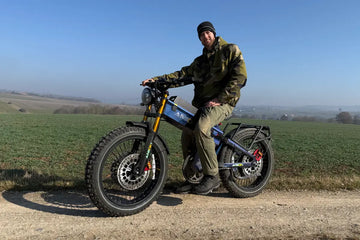 A Caucasian man in a camo jacket sits on a blue Aniioki A9 electric bike on a dirt road, showcasing the disc brake system to prevent e-bike brakes squeaking after rain