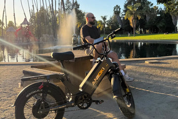 A Caucasian man relaxing near a fountain in a sunny outdoor park beside a black Aniioki AQ177 48V long-range electric bike, showcasing modern e-bike battery types and design.