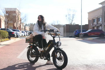 A young Caucasian woman in a white jacket riding a black Aniioki AQ177 Pro Max ebike through a suburban shopping center parking lot during the day.