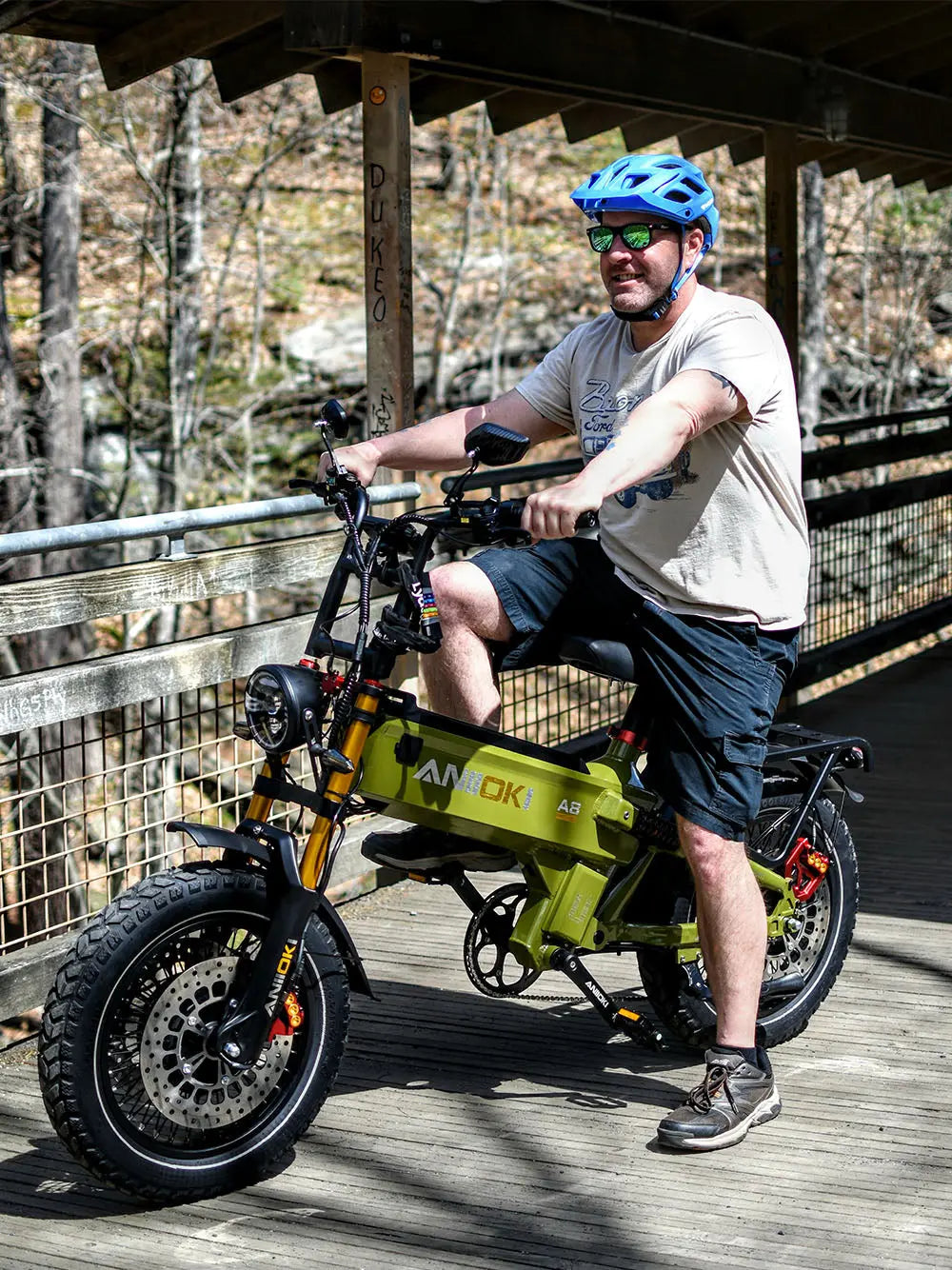 A middle-aged white man riding an army green Aniioki A8 Pro Max 2025 on a wooden bridge, a durable and affordable ebike for daily commuting and trail riding.