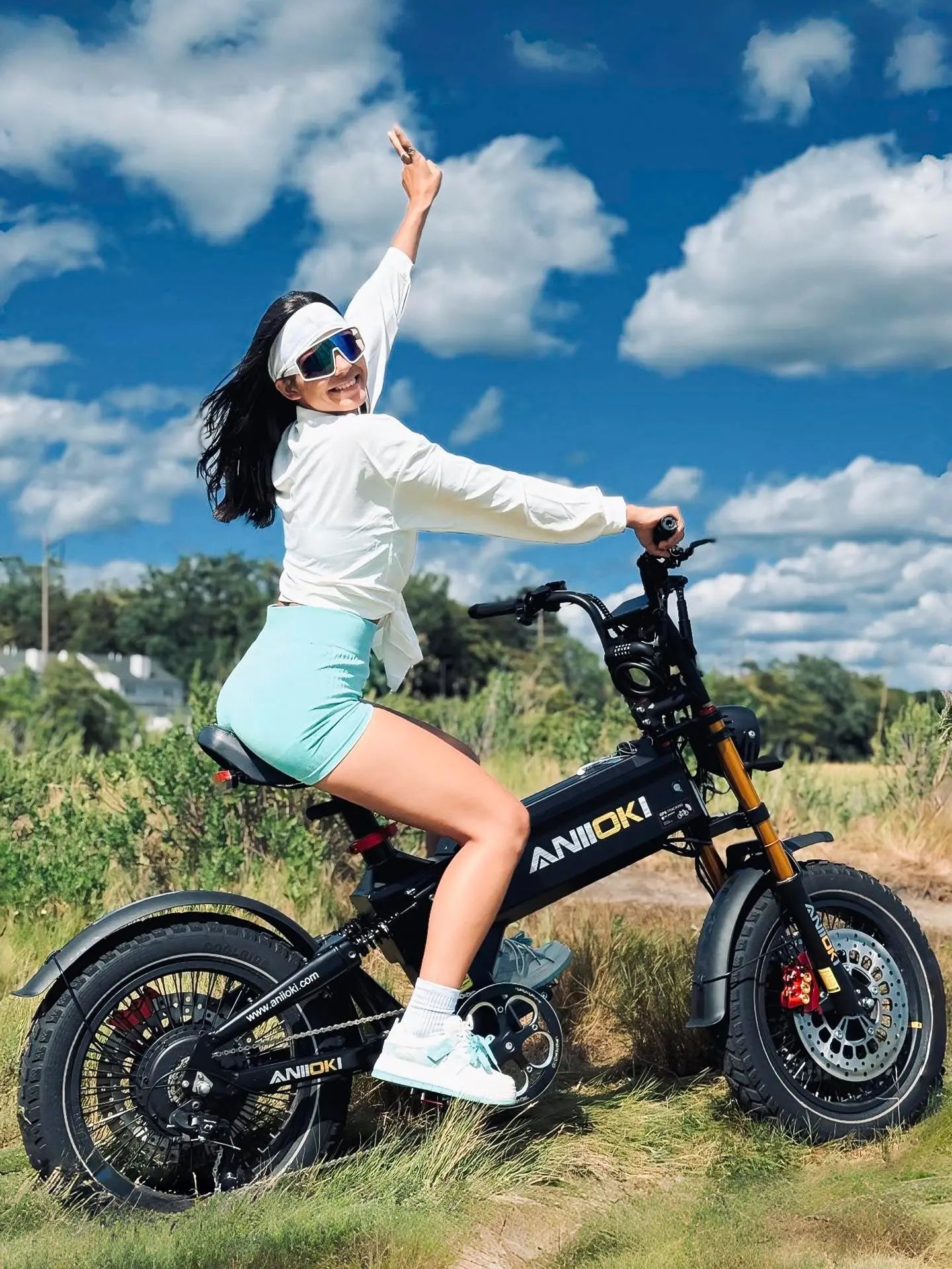 A happy Asian woman riding a matte black Aniioki A8 Pro Max 60V dual motor ebike under a blue sky, showcasing the stylish design of this electric dual motor bike.
