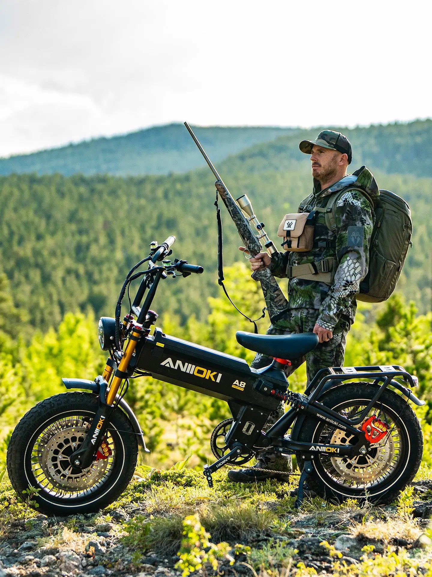  A white man in camouflage gear with a rifle standing next to a black Aniioki A8 Pro Max dual motor, perfect 6000w dual motor electric bike for outdoor hunting and exploration.