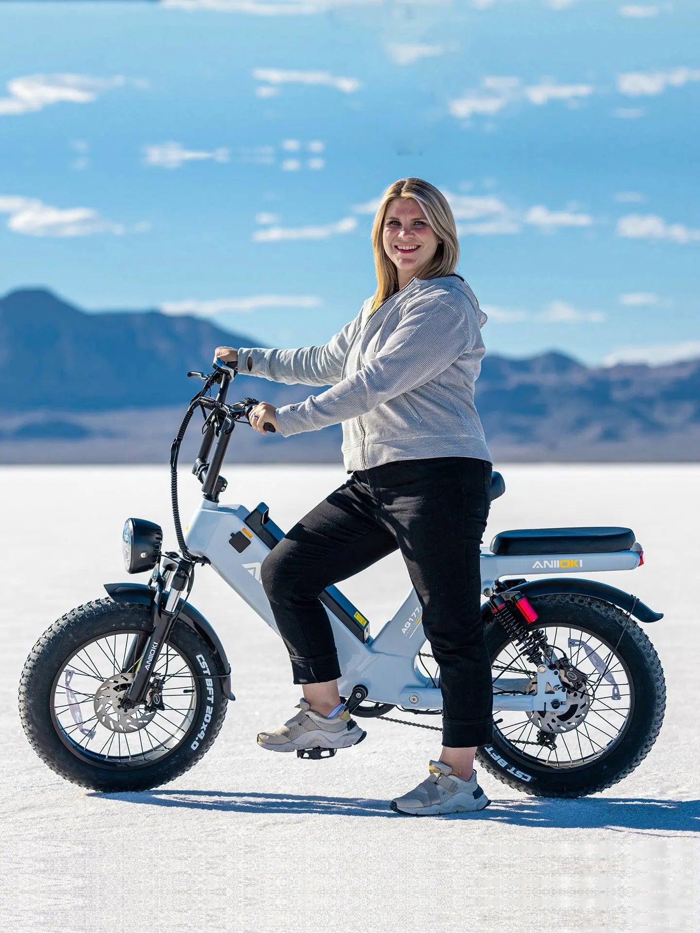 A woman posing on a gray Aniioki AQ177 Pro Max, a step-thru ebike with electric motorcycle range and fat tires for stable riding on flat terrain.